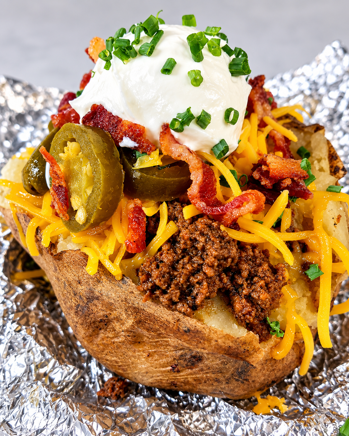 Loaded baked potato with taco toppings — seasoned ground beef, shredded cheddar, crispy bacon, jalapeños, sour cream and chives on foil